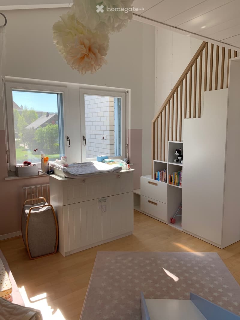 A nursery with a changing table, white cabinets, and a bookshelf. A toy and books are on the shelf. A chair and a rug are on the floor. A box and bottles are on the window sill. A staircase is on the right side. A ceiling decoration is hanging from the ce