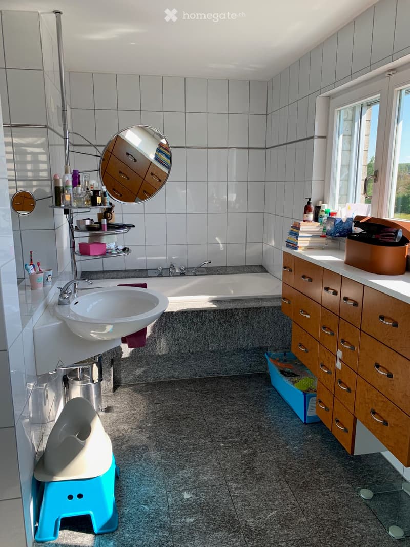 Bathroom with white tiles, a sink, mirror, bathtub, and wooden cabinets. A toilet seat cover and toothbrushes are on the wall.