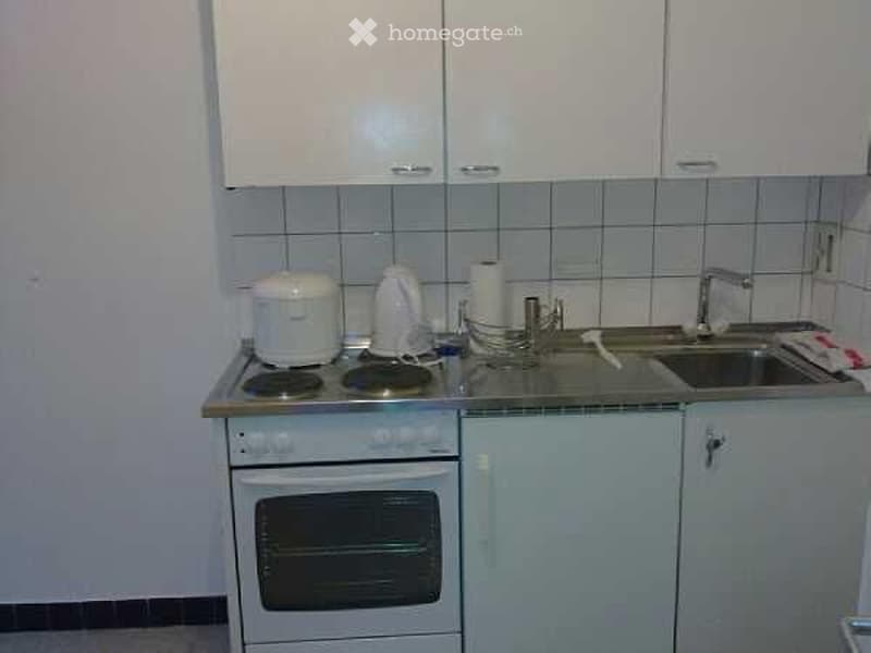A compact kitchen with white cabinets, a small electric stove, a sink with a faucet, and various small appliances on the counter.