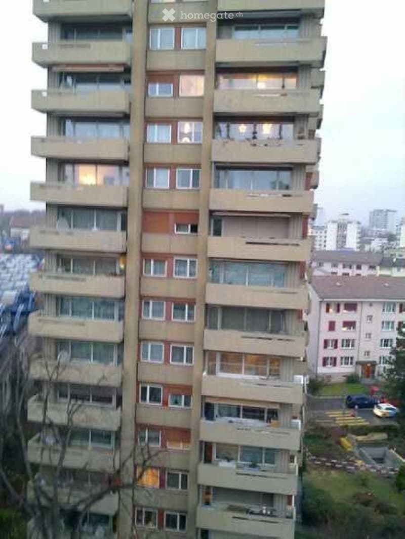 A tall, multi-story residential building with numerous windows and balconies, surrounded by other buildings and a parking area.