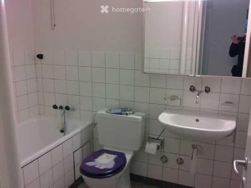 A small bathroom with white tiled walls and floor, featuring a toilet with a purple seat cover, a sink, a bathtub, and a mirror above the sink.