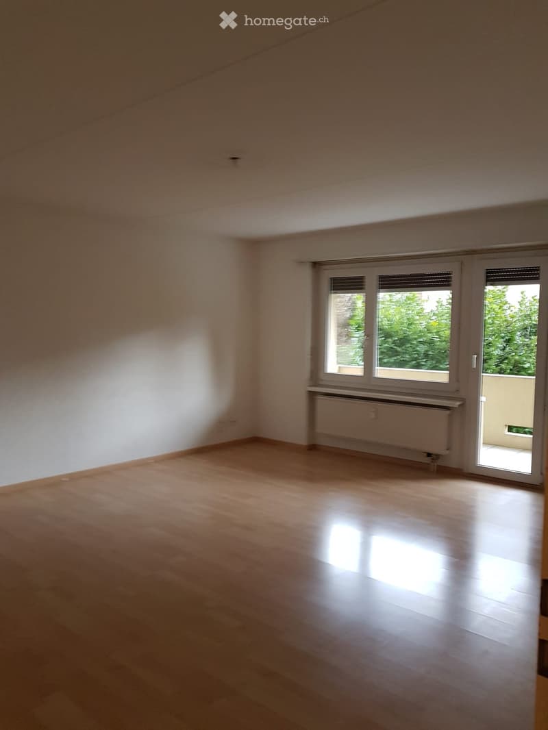 Empty room with wooden flooring, white walls, large windows, and a sliding glass door leading to a balcony.