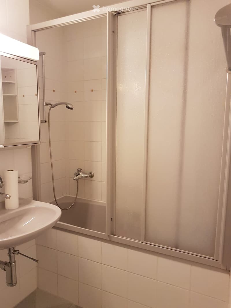 Modern bathroom with white tiles, a sink, a bathtub with a glass shower enclosure, and a mirror with built-in shelves.