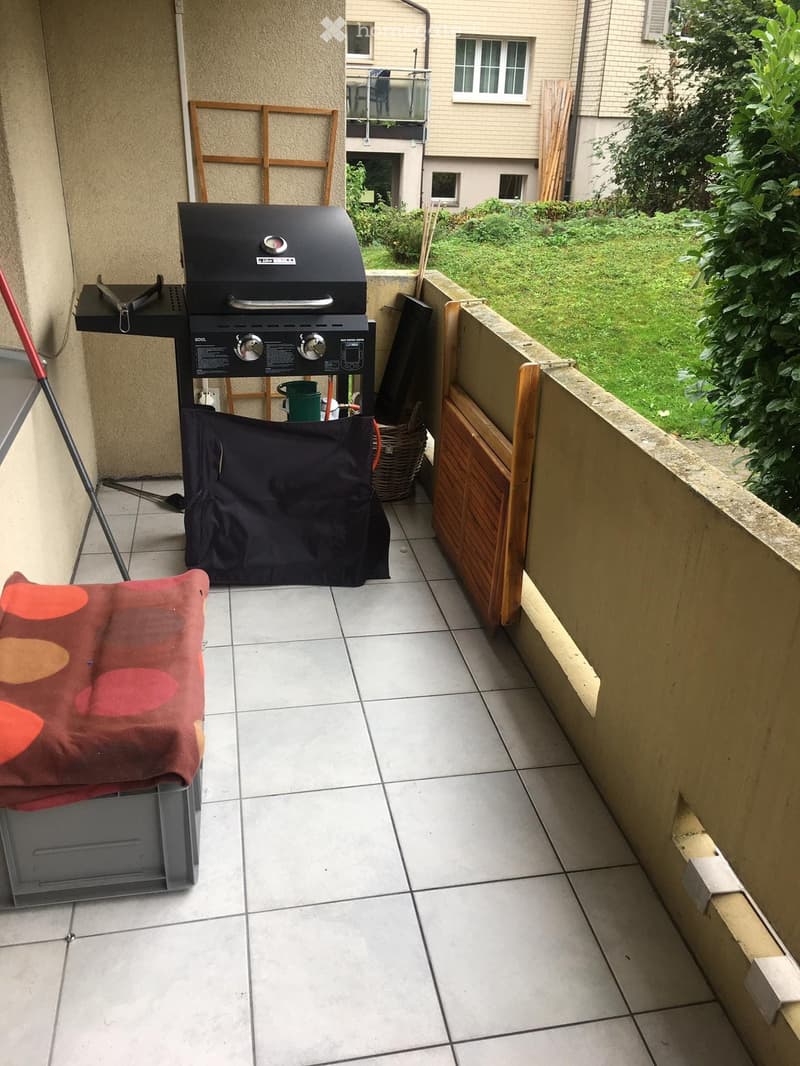 Balcony with tiled floor, barbecue grill, covered grill, small wooden cabinet, and a red cushion on a grey storage box.