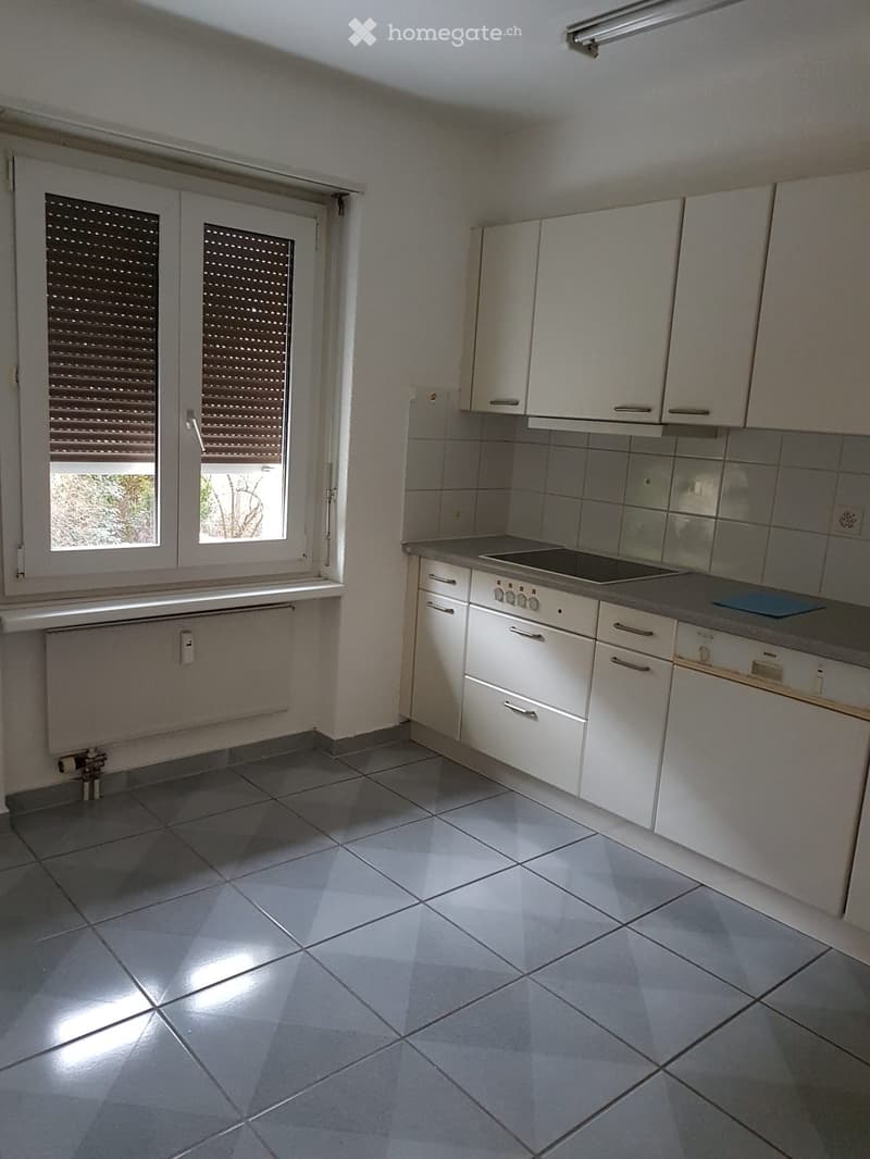 Empty kitchen with white cabinets, tiled floor, and a window with blinds. No appliances or furniture are present.