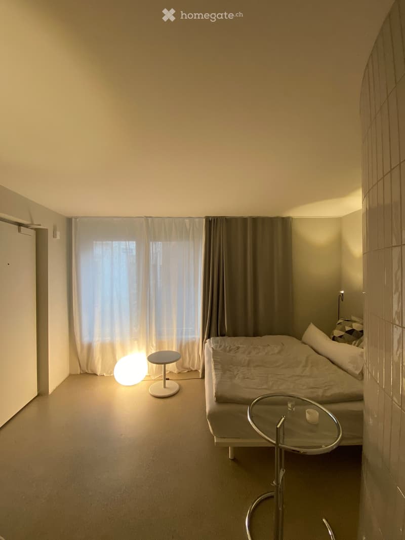 A small, modern bedroom with a bed, a round glass table, a stool, and a spherical lamp near the window.