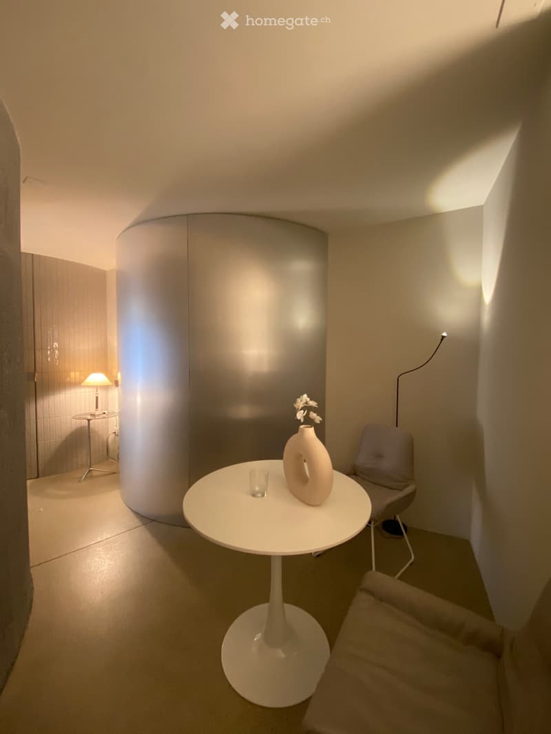 Minimalist room with a white round table, vase, cup, chair, and lamp. Features a curved wall and soft lighting.