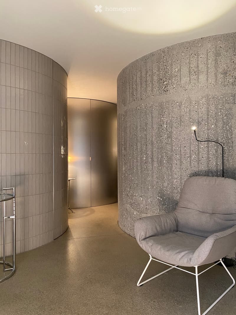 Modern interior with a grey upholstered chair, a sleek round glass table, and a textured wall in a contemporary design.