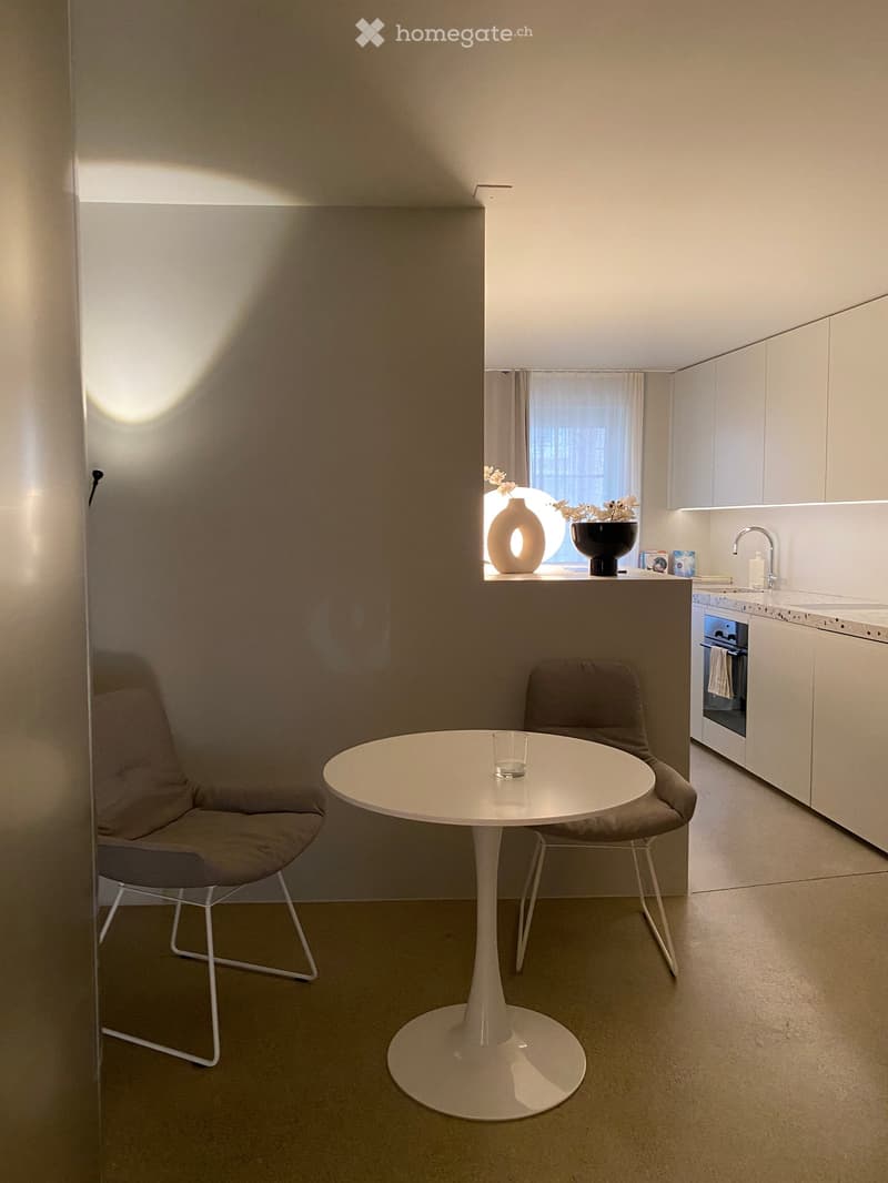 A modern, minimalist kitchen and dining area with a white table, two chairs, a vase on the windowsill, and sleek white cabinets.