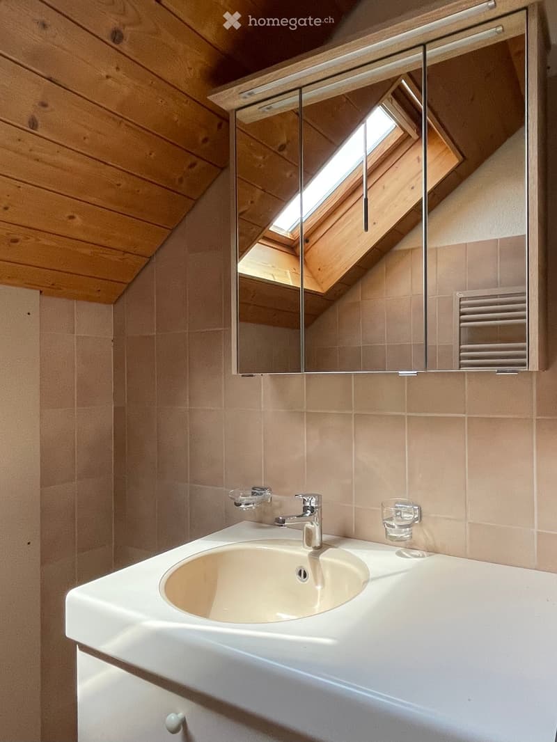 Modern bathroom with a white sink, chrome faucet, large mirror, and a skylight window above the mirror. Beige tiles and wooden slanted ceiling.