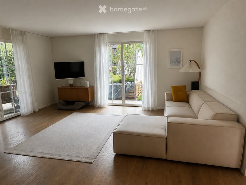 Modern living room with light-colored sofa, coffee table, television, floor lamp, and large windows leading to a balcony.