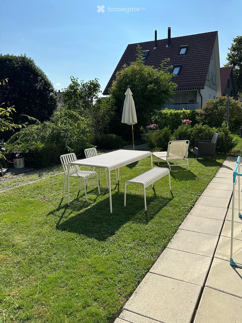 Garden with a white table, chairs, umbrella, and a bench on a grassy area next to a house.