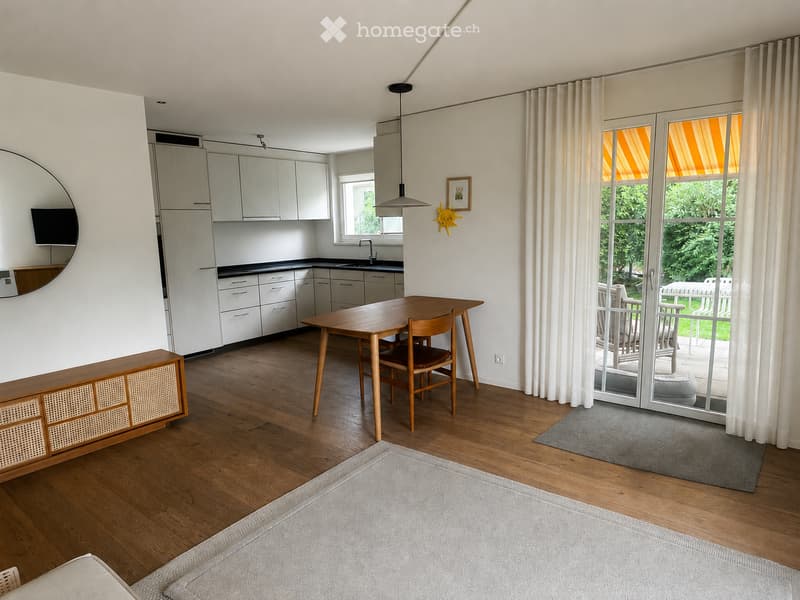 Open-plan living and dining area with wooden flooring, white kitchen cabinets, a dining table with chairs, a sofa, and a large window with white curtains leading to a balcony.
