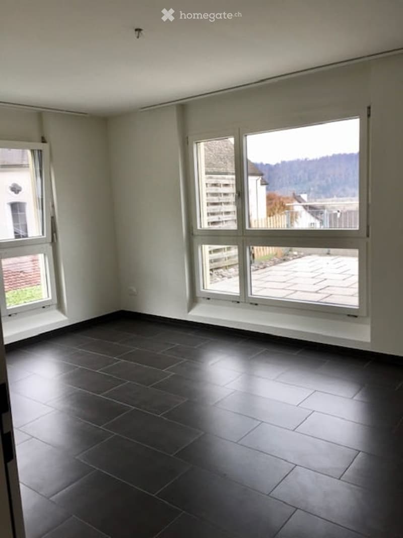 Empty room with dark tiled flooring, white walls, and large windows offering a view of the outdoors.