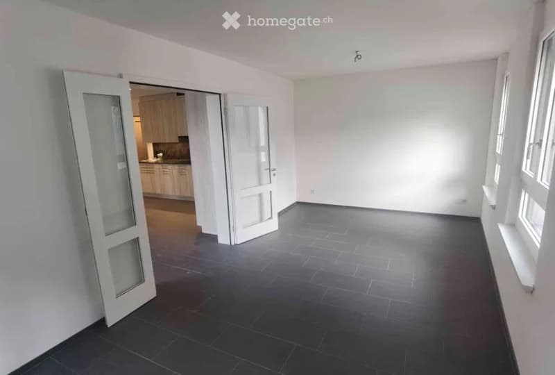 Empty room with dark tiled flooring, white walls, and large windows. An open doorway leads to a kitchen area with cabinets and a countertop.