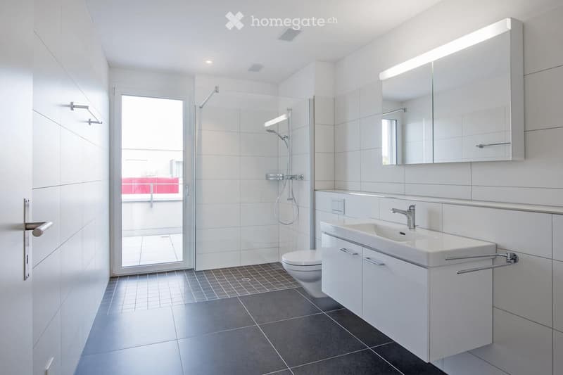 Modern bathroom with white tiles, a large mirror, a sink with cabinet, a toilet, and a glass-enclosed shower. The room features sleek, contemporary design with built-in storage and bright lighting.