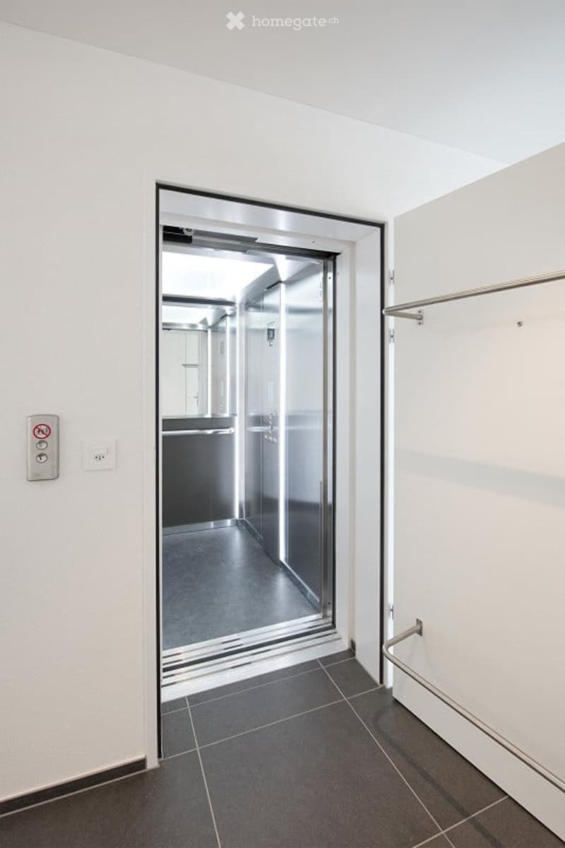 Modern hallway with a glass elevator door, sleek metallic frame, and a clean, minimalist design. The floor is tiled in dark grey, and the walls are white.