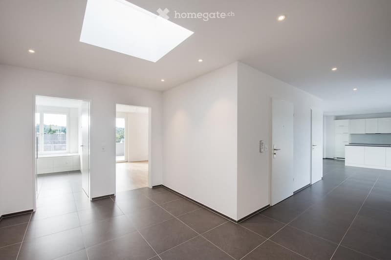 Bright, empty room with white walls, dark tiled floor, and recessed lighting. Features include a skylight, several doors leading to other rooms, and a view of a kitchen area with white cabinets.