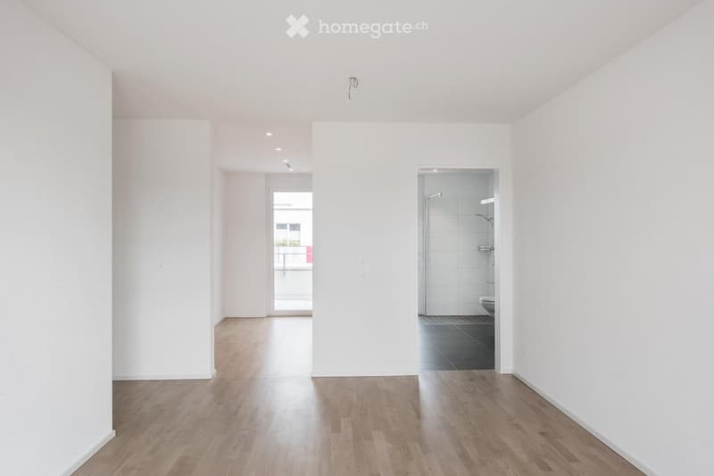 Empty hallway with light wooden flooring, white walls, and a view of a bathroom and balcony area.