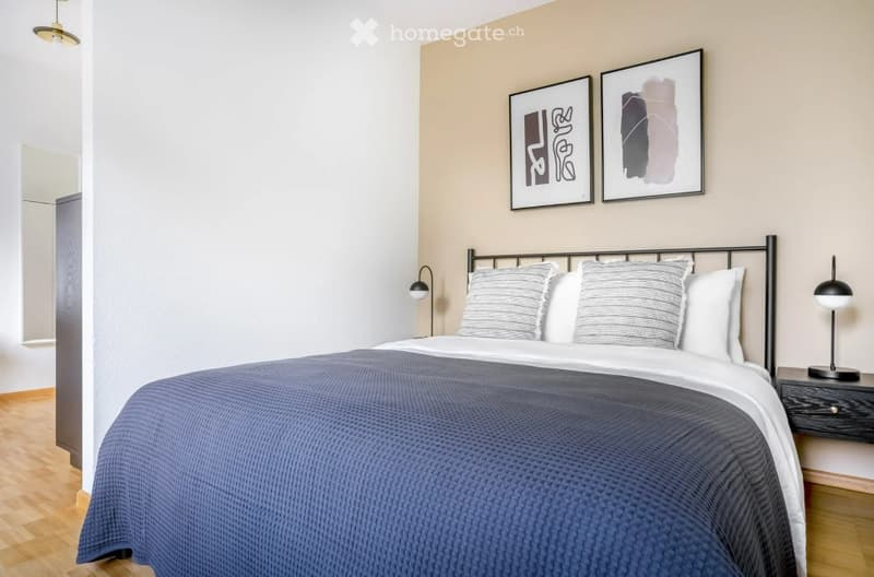 Modern bedroom with a neatly made bed, nightstand, lamp, and two picture frames on the wall.