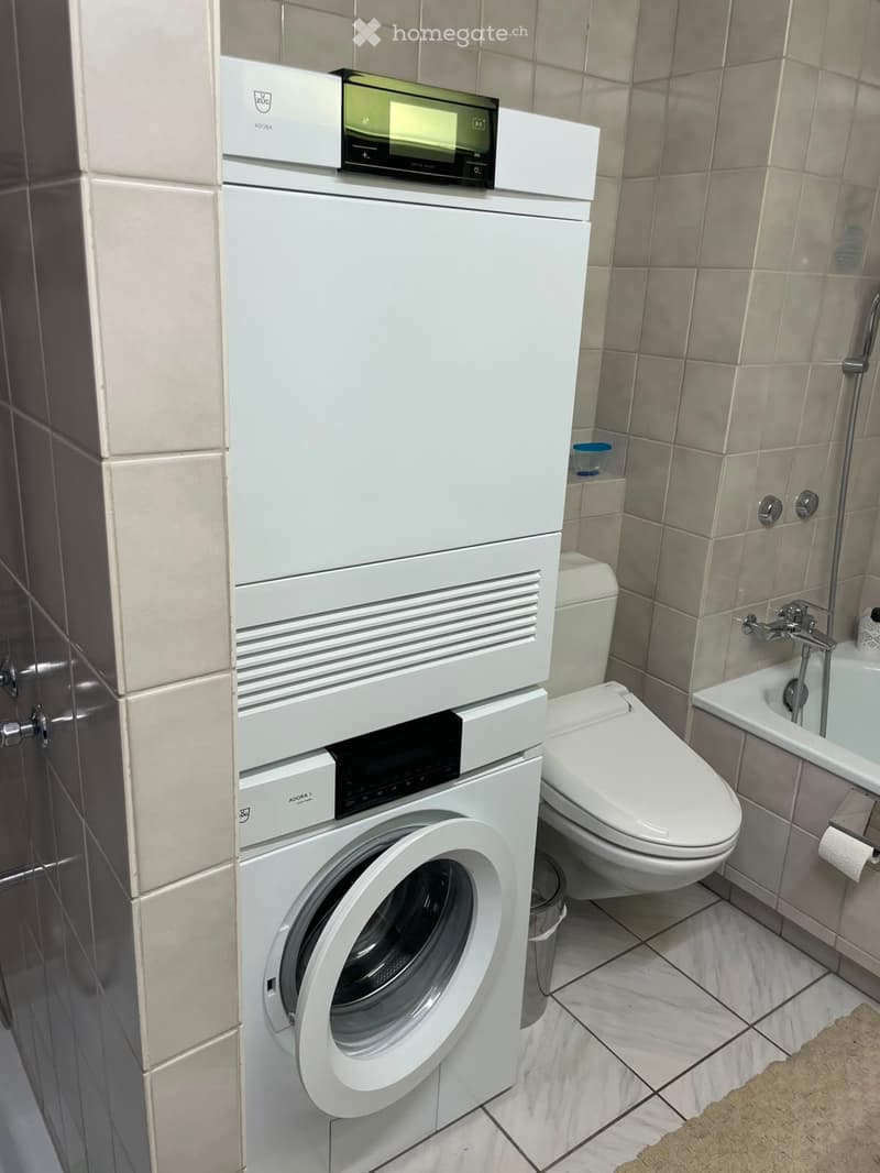 Modern bathroom with a white washing machine, toilet, and bathtub, featuring tiled walls and a beige floor.