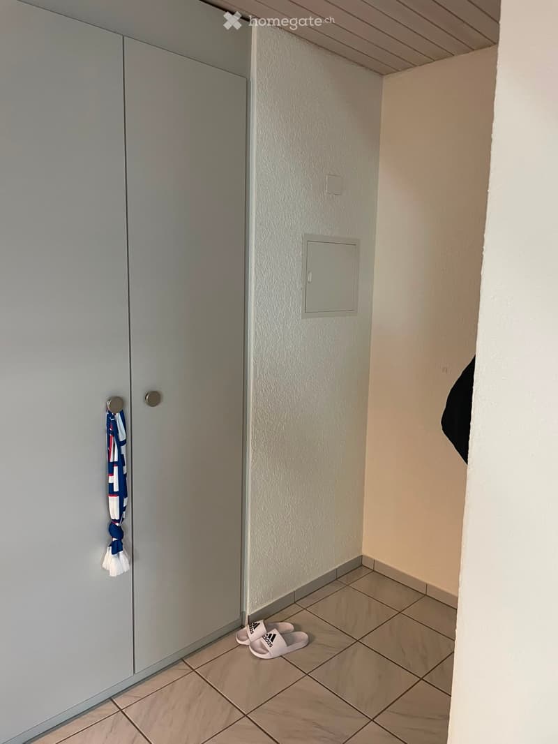 A narrow hallway with tiled flooring, white walls, and a closed door on the left. A pair of white Adidas slides are placed near the door, and a small decorative item hangs on the wall.