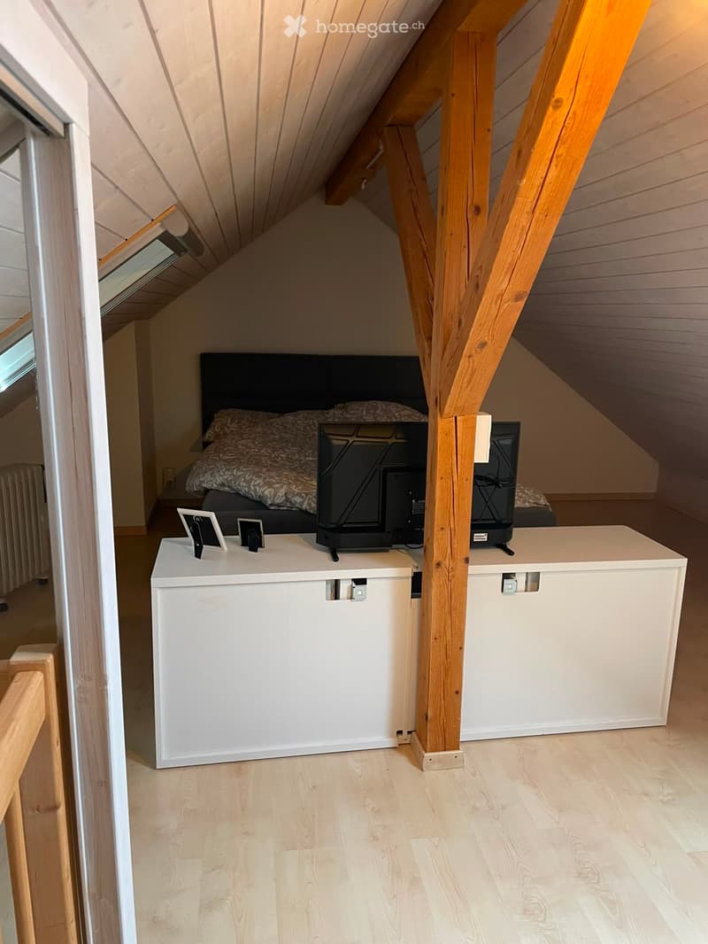 Cozy attic bedroom with wooden beams, slanted ceiling, bed with headboard and pillows, small table with screen and picture frames.