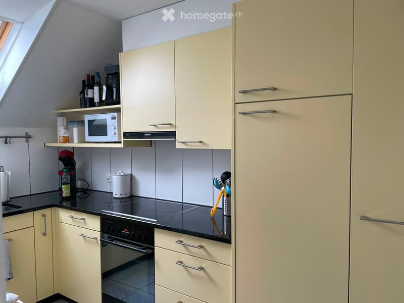 Modern kitchen with light-colored cabinets, stainless steel handles, and a black countertop. Appliances include a microwave, oven, and coffee maker. Shelves hold bottles and other kitchen items.
