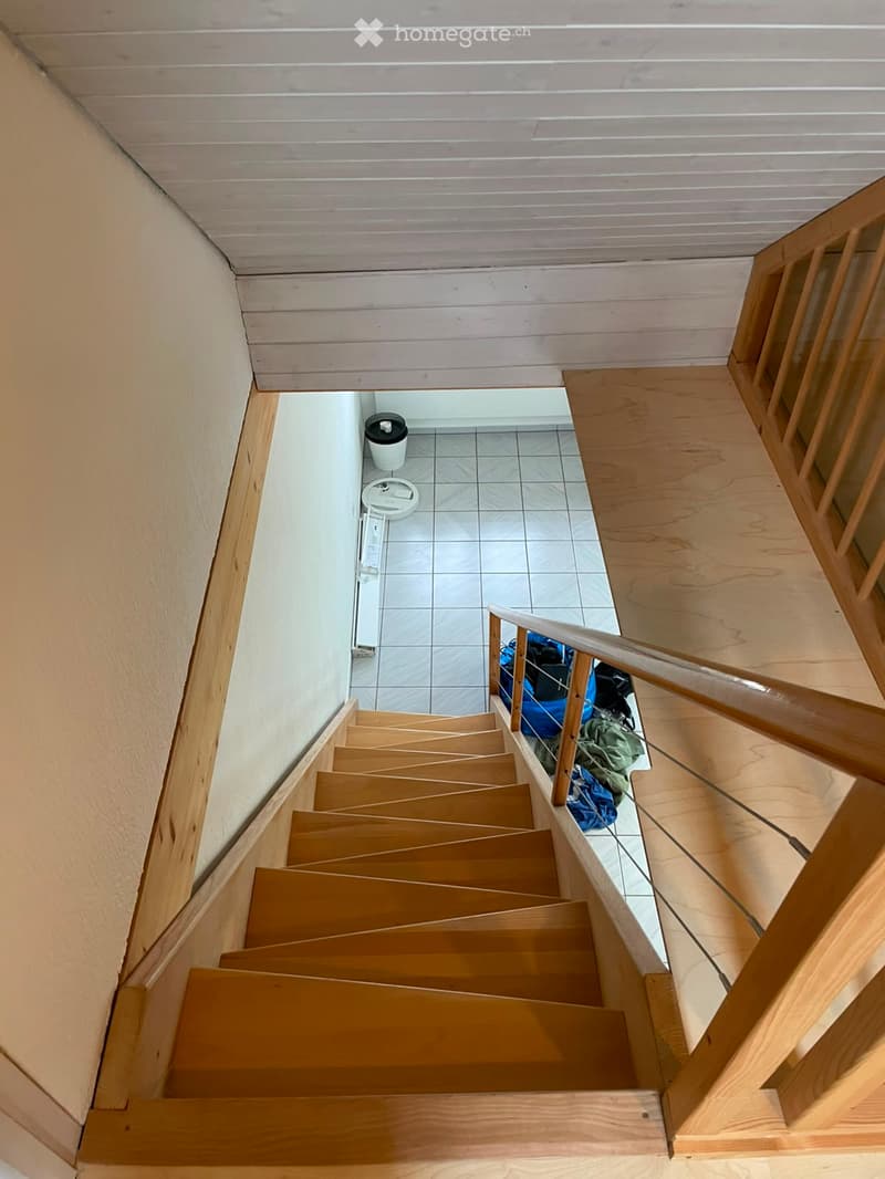 Wooden staircase with a handrail leading to a tiled area below, featuring a small trash bin and some bags on the floor.