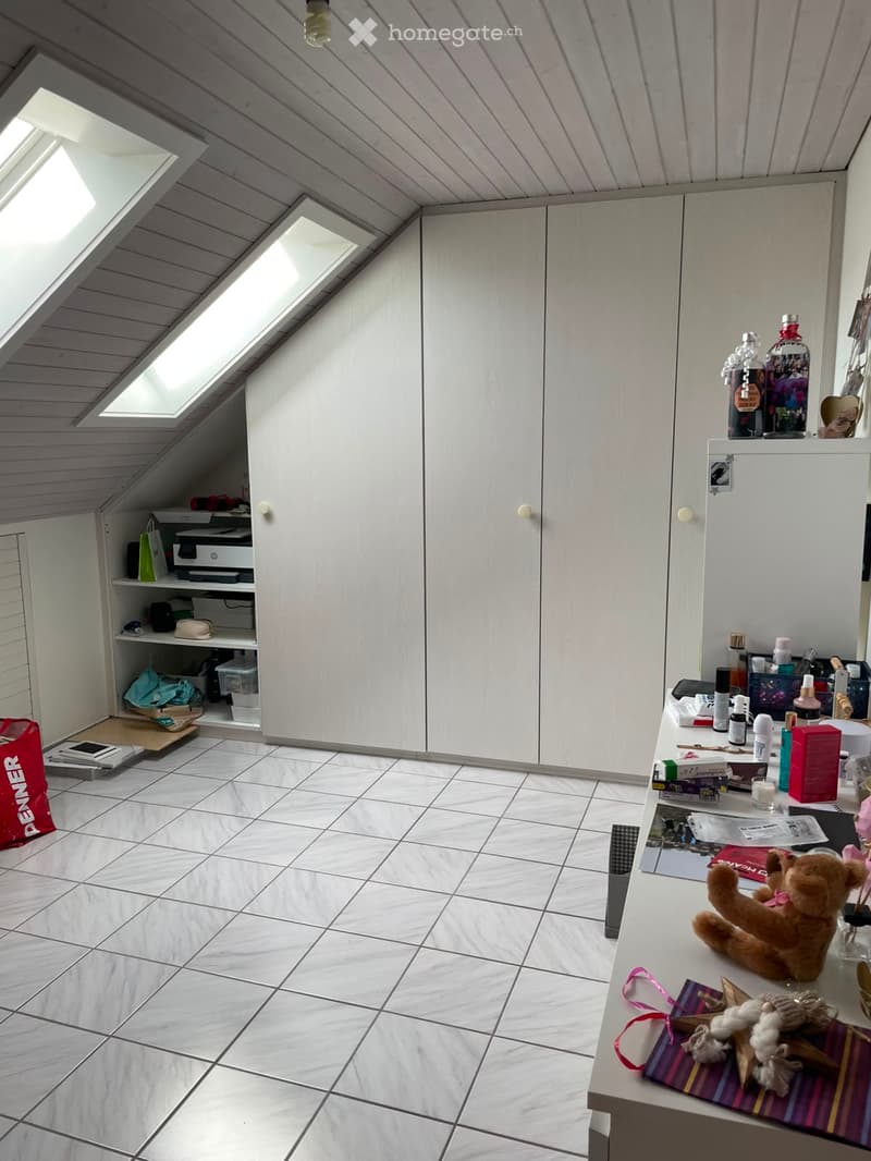 A well-lit attic room with white walls, tiled floor, and built-in wardrobes. The room features a small table with a teddy bear, bottles, and other items, along with a printer and shelves in the corner.