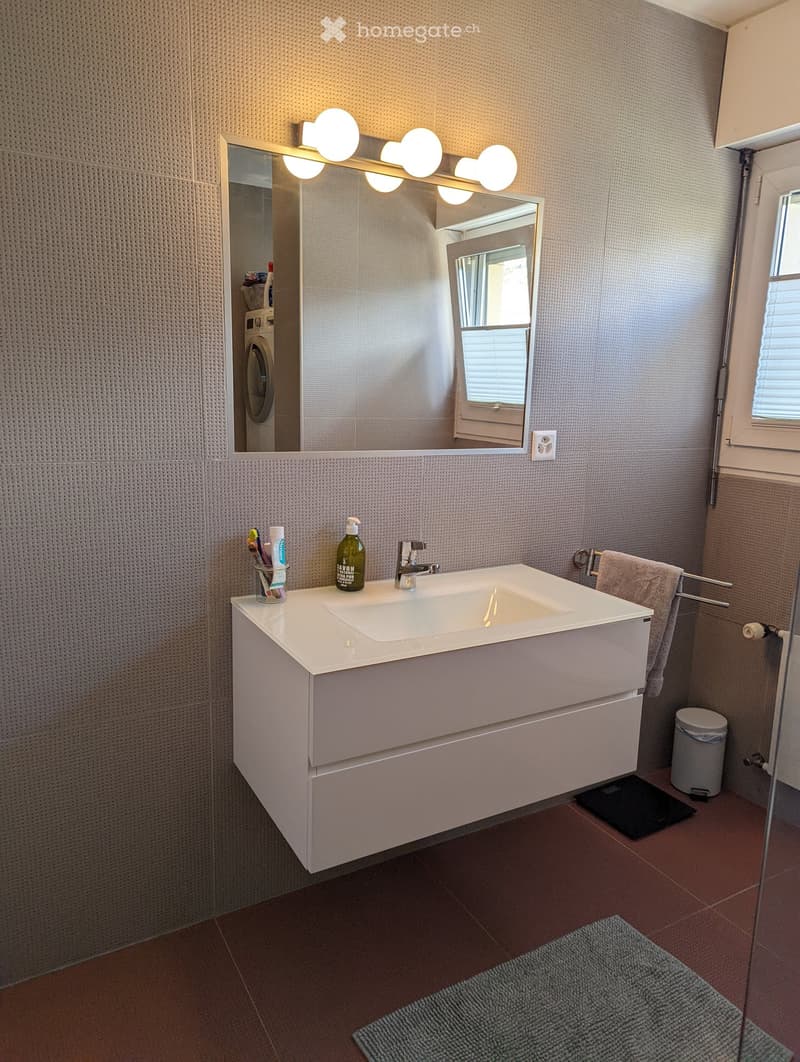 white vanity with sink, mirror, faucet, toiletries, trash bin, towel rack, towel, brown tile flooring, glass door, wall outlet, window