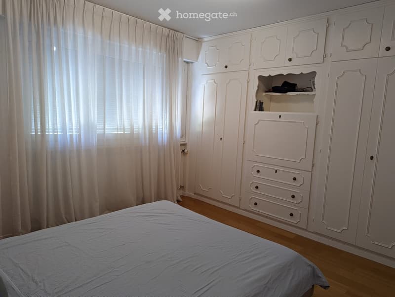 Wooden floor, white walls, large white closet, bed with a gray bedsheet, white curtain