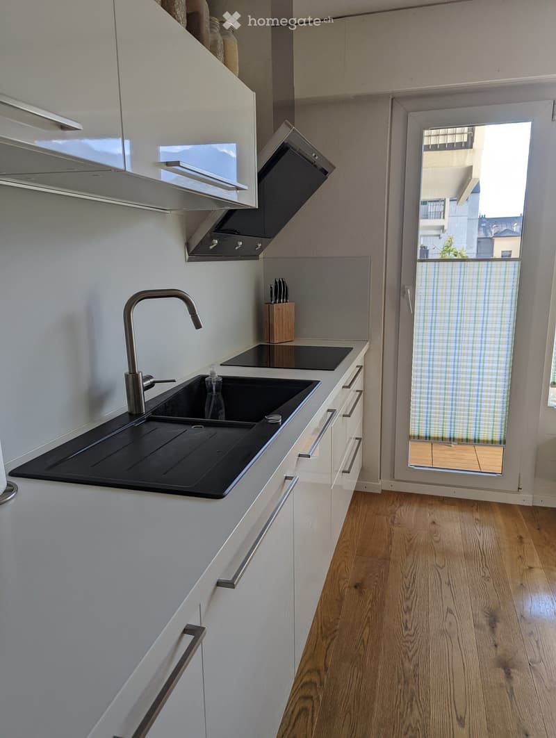 modern design with white cabinets, wooden floors, black kitchen island with granite countertop, under-cabinet lighting, stainless steel gas stove and sink, glass door leading to balcony