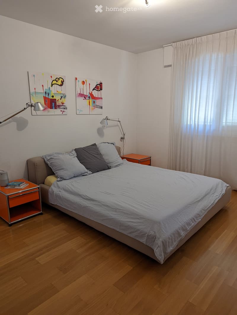 Modern bedroom with wooden floor, double bed with light grey sheets, two nightstands, lamps on either side of the bed, two paintings on the wall, and white curtains covering the window. A small orange object is on the nightstand, and books are on top of t