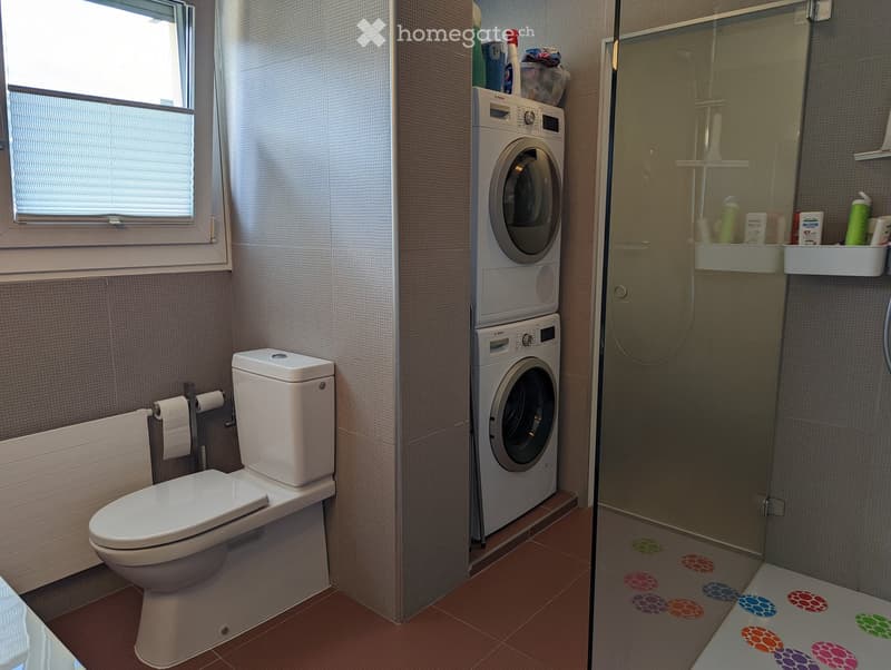 Compact bathroom with white toilet, stacked washer and dryer, white shelves, tiled floor, glass shower, white radiator, and white blinds.