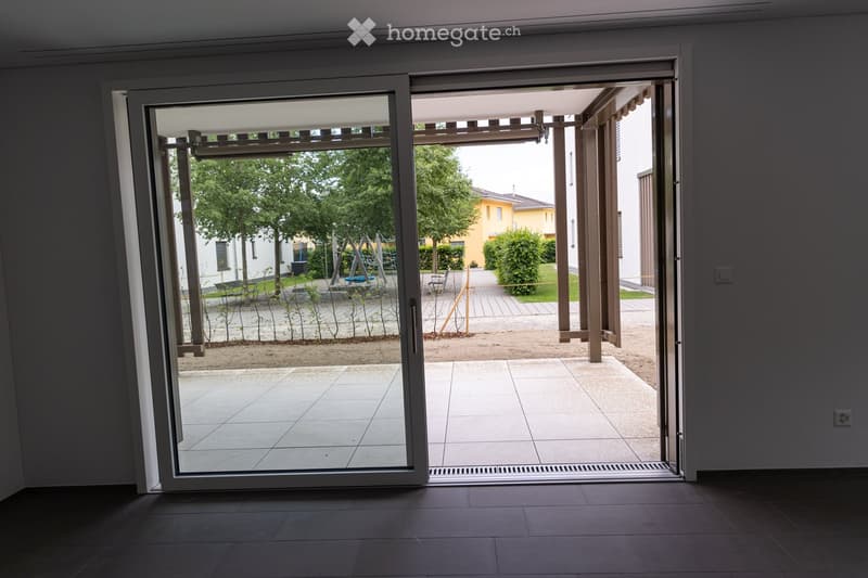 An empty room with dark tiled flooring and large sliding glass doors leading to a tiled patio area. The patio features a gravel pathway, small plants, and a view of a landscaped garden with trees and a yellow building in the background.