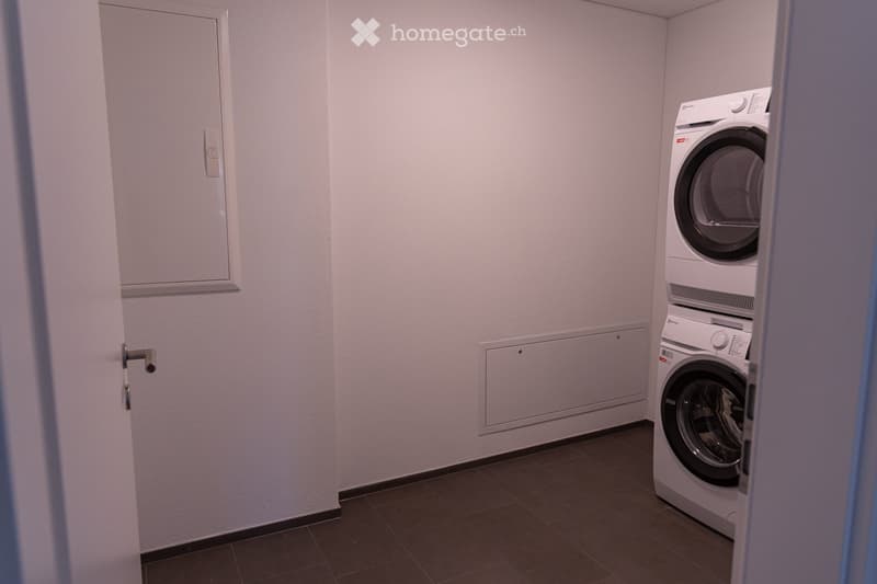 A small, well-lit room with a washing machine and dryer stacked vertically, light-colored walls, and a tiled floor.