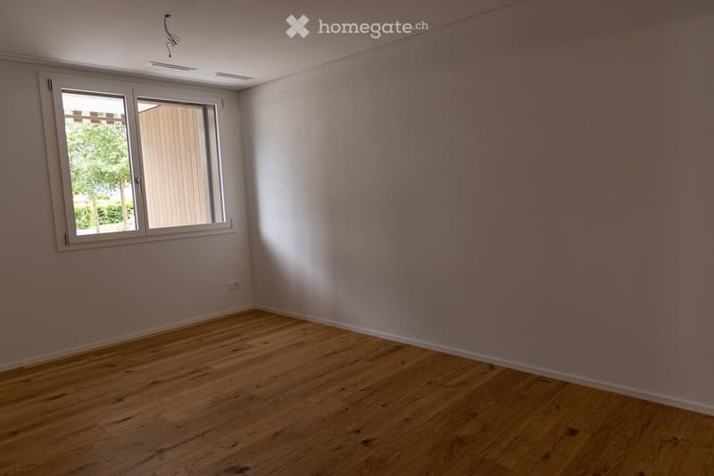 Empty room with wooden flooring and white walls, featuring a large window with white frames and blinds, and a visible electrical outlet on the wall.