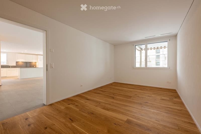 Empty room with wooden flooring, white walls, and a large window. The adjacent kitchen area is visible through an open doorway.