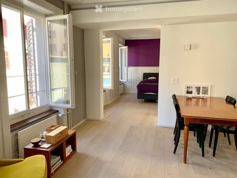 Bright, open-plan living and dining area with wooden flooring, a yellow sofa, a wooden dining table with chairs, a small shelf with books and a bowl, and a bedroom with a purple accent wall and a bed.
