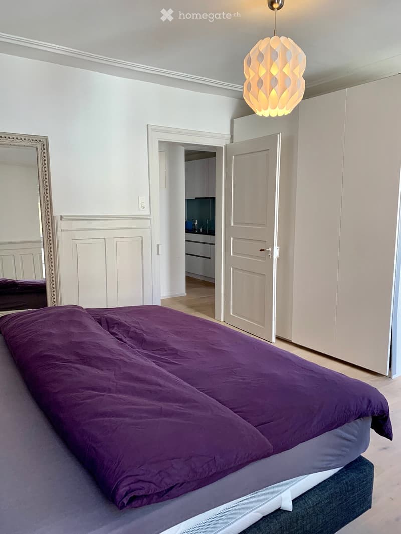 Modern bedroom with a large bed covered in purple bedding, a decorative mirror, and a built-in wardrobe. The room features light wooden flooring and white walls.