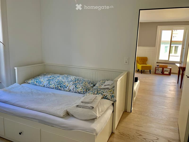 Cozy bedroom with two single beds, a yellow armchair, a small table, and a stool near a large window with a view.