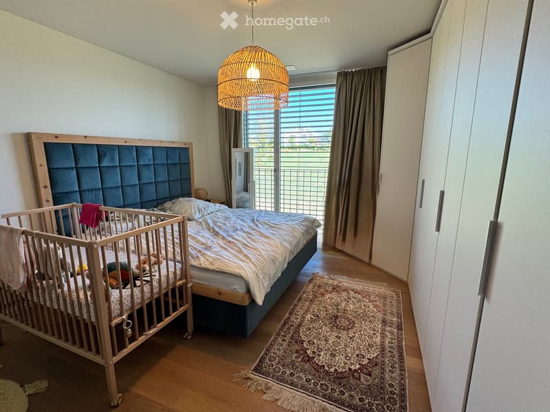 Bright bedroom with a large double bed, a baby crib, a patterned rug, and a modern woven pendant light. The room features wooden flooring, white walls, and a large window with blinds.