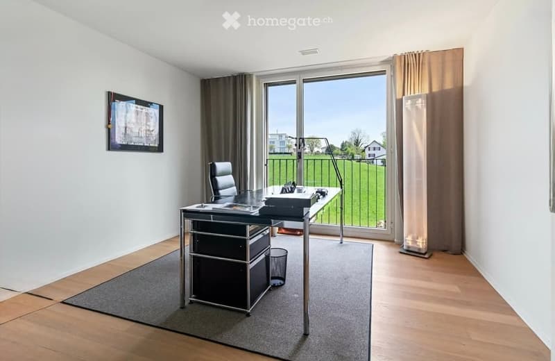 Modern office space with wooden flooring, large window offering a view, desk, chair, screen, lamp, and picture frame on the wall.
