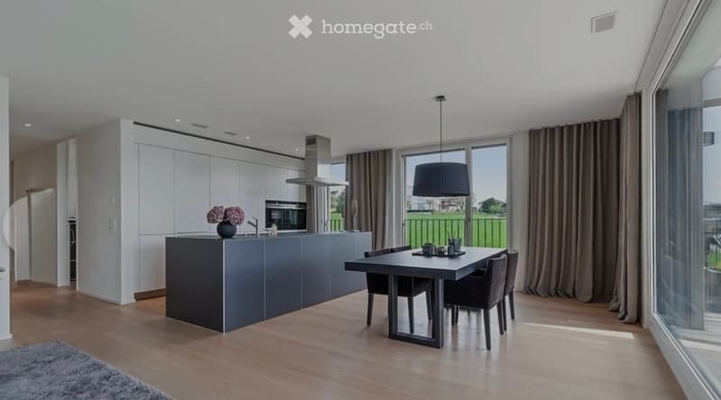 Modern kitchen and dining area with a grey island, black dining table, four chairs, pendant light, large windows with curtains, and a vase with flowers on the island.