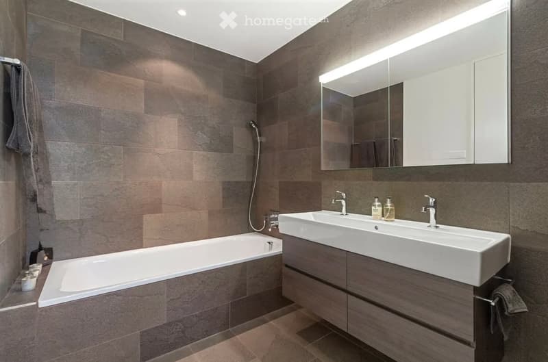 Modern bathroom with dark tiled walls, a bathtub, a double sink with modern faucets, a large mirror with built-in lighting, and a towel hanging on the side.