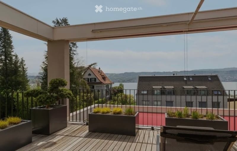 Modern balcony with wooden decking, black metal railing, potted plants, and scenic view of lake and surrounding landscape.
