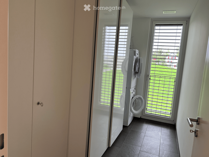 A narrow hallway with a washing machine and dryer placed against the wall, large windows with blinds, and a door leading outside.