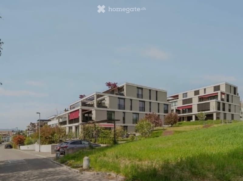 Modern multi-story residential building with balconies, surrounded by greenery and a paved pathway.