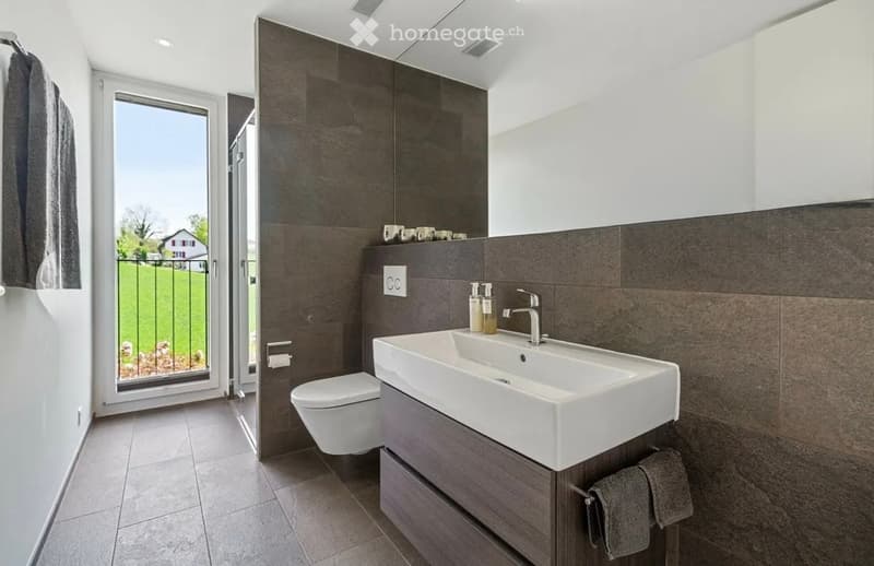 Modern bathroom with a white sink, toilet, dark tiled wall, and a large window offering a view of a green lawn and house.